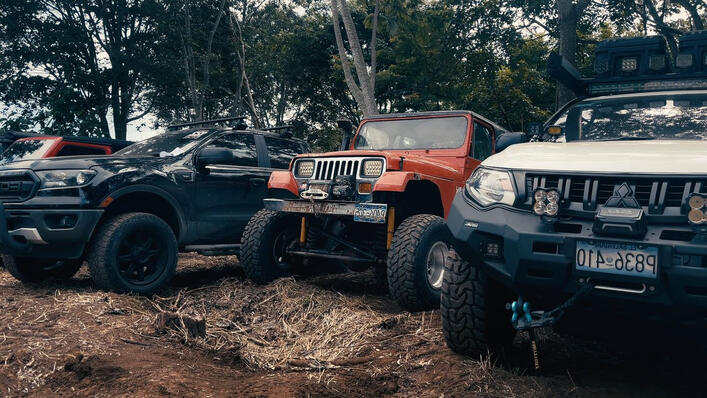 Collection of modified 4x4 off-road vehicles, including a Jeep Wrangler, Ford Ranger Raptor, and Mitsubishi pickup, parked before an off-road adventure event in a forest environment.