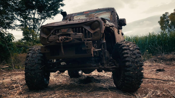 A muddy, off-road vehicle with large tires parked on a dirt path, surrounded by trees and tall grass under a cloudy sky.